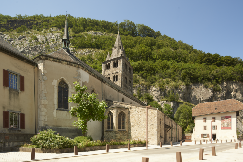 Historic stone church with a bell tower surrounded by green hills on a sunny day in Saint-Ursanne, Switzerland.