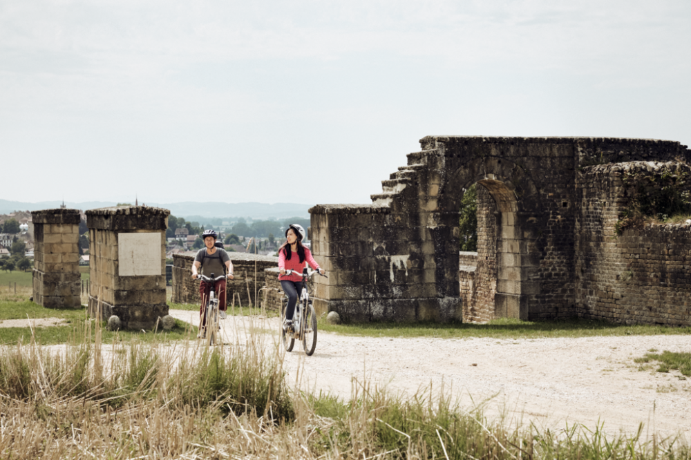 Cyclists riding along the ancient Via Romana surrounded by historic ruins and green landscape on a sunny day.