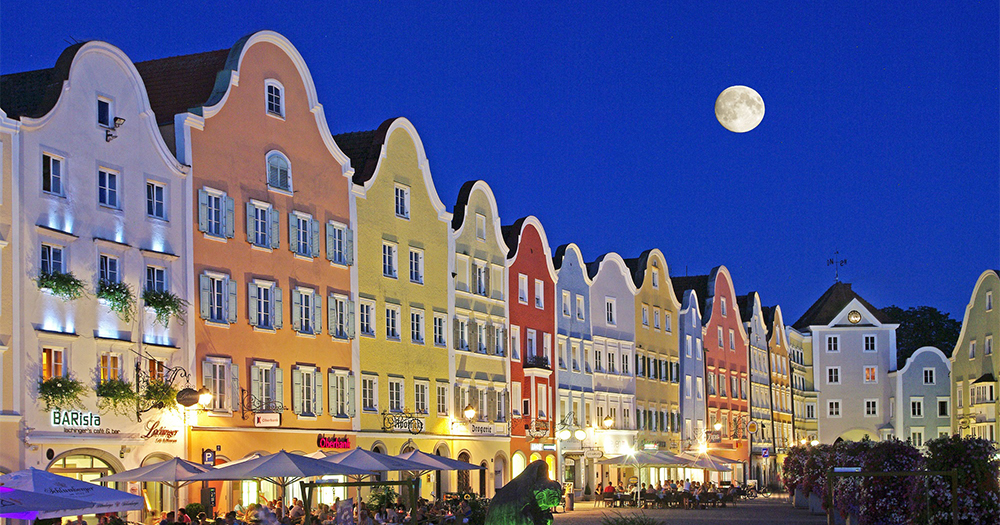 Lively plaza in Schärding at night, featuring colorful Baroque architecture and a glowing full moon.