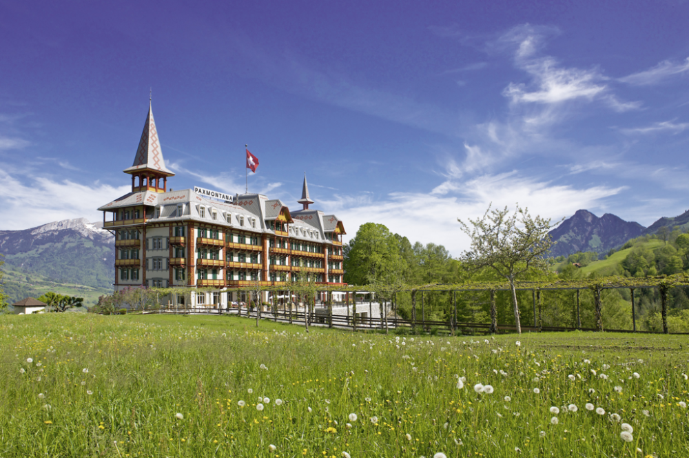 View of a hotel in the green landscape of Kerns village, with a Swiss flag and dandelions under a sunny sky with light clouds.