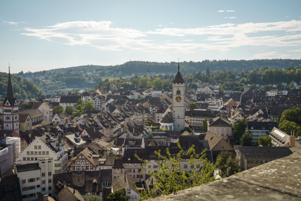 View of the old town of Schaffhausen on a sunny day, with white buildings, a clock tower, a church, and green hills in the background.
