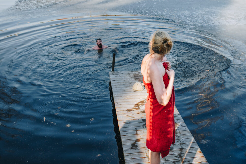 Woman in red towel standing on a wooden dock by the water with a person swimming.